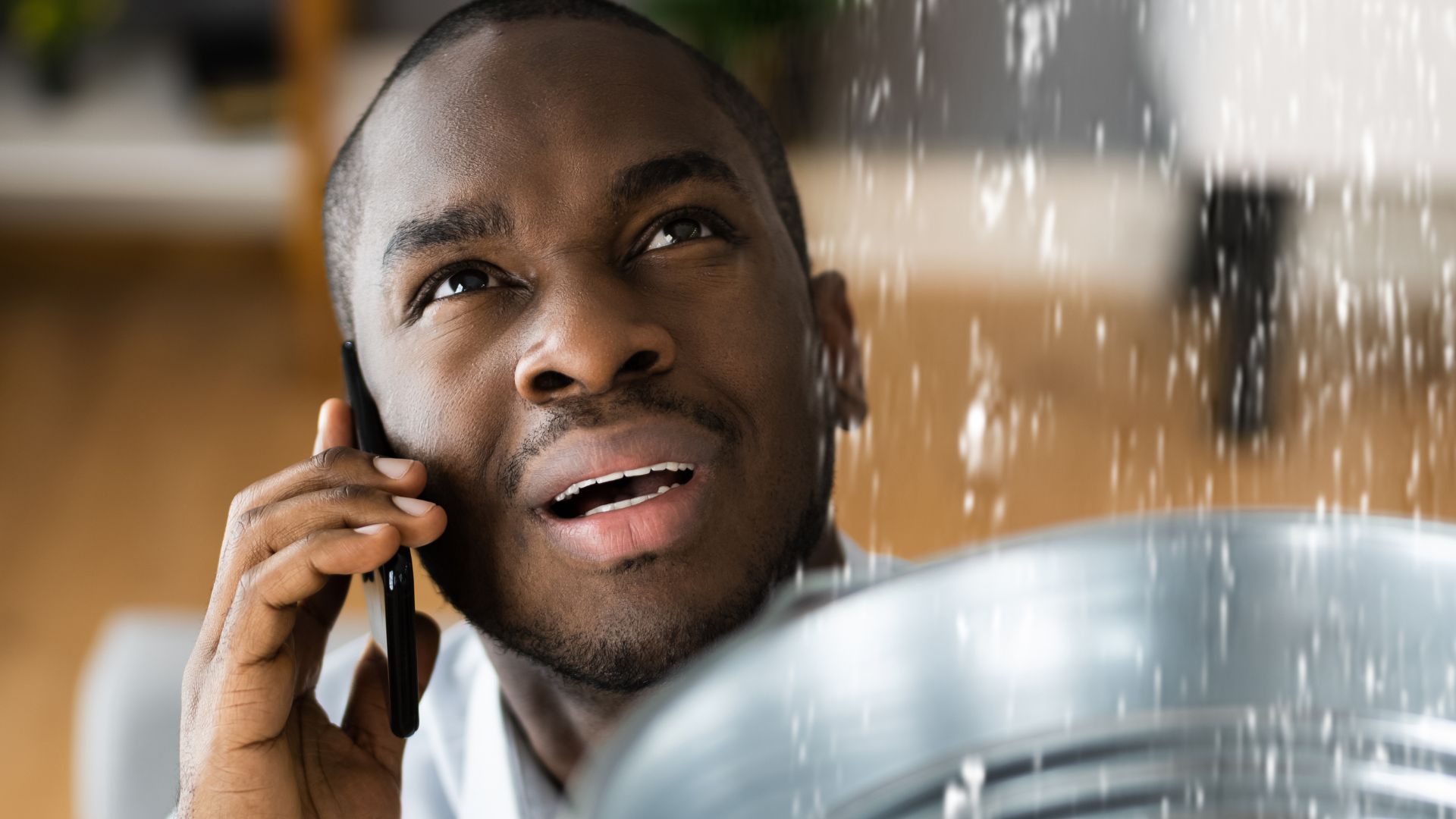 upset man on phone, holding bucket to catch roof water